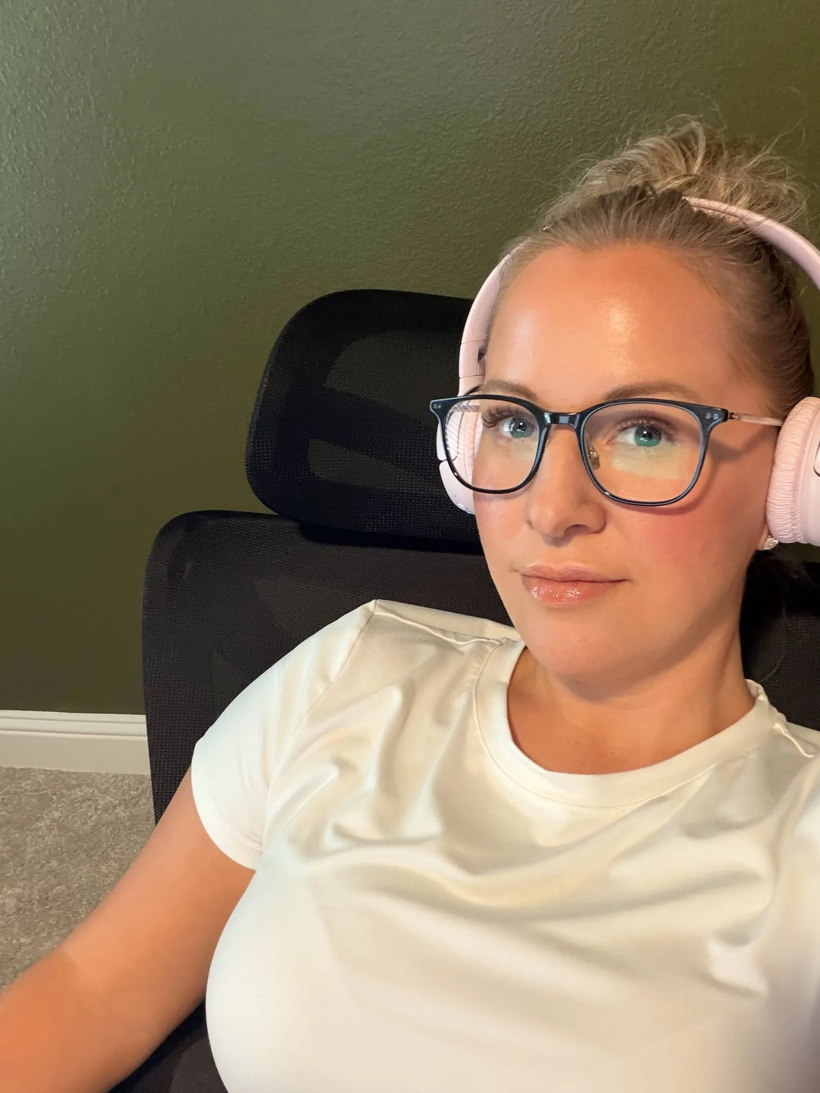 Tegan, wearing pale pink headphones and dark-framed glasses, looking toward the camera from a black mesh desk chair against an olive green wall.